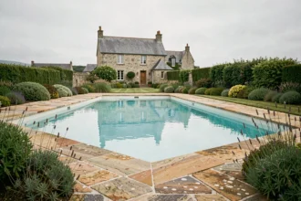 Piscine extérieure aménagée dans un jardin breton avec dallage en pierre naturelle et maison en granit en arrière-plan, illustrant le marché des constructeurs de piscines dans le Morbihan